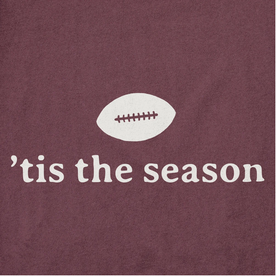 A close-up of a maroon fabric surface with a white football graphic and the phrase "'tis the season" in white lowercase text.