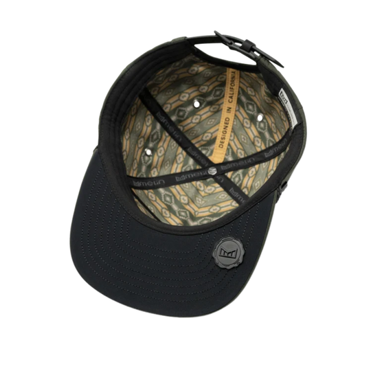 A black structured baseball cap viewed from the front, showing a patterned olive green and tan interior with black elastic straps and a small circular black logo patch on the front brim.