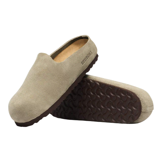 A pair of slip-on clog-style slippers in light beige suede, with one shoe standing upright and the other lying on its side to show the dark brown textured sole. The brand name 'Birkenstock' is printed on the side of the upper.