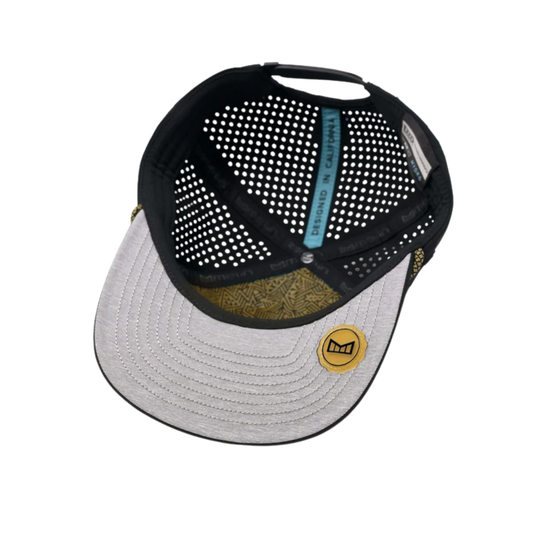 A trucker-style baseball cap shown from a top-down angle on a white background. The cap features a black mesh back, a light heather gray brim, a gold patterned band, and a small gold circular patch on the brim.