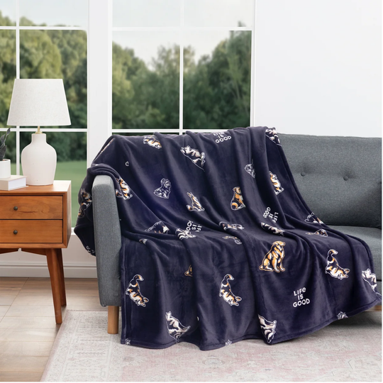 A navy blue fleece throw blanket with a playful dog print and the words 'Life is  good' draped over a gray sofa in a bright living room setting.