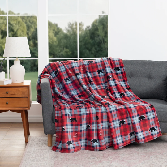 A red and blue plaid throw blanket with black bear motifs is draped over a gray sofa in a bright living room setting.