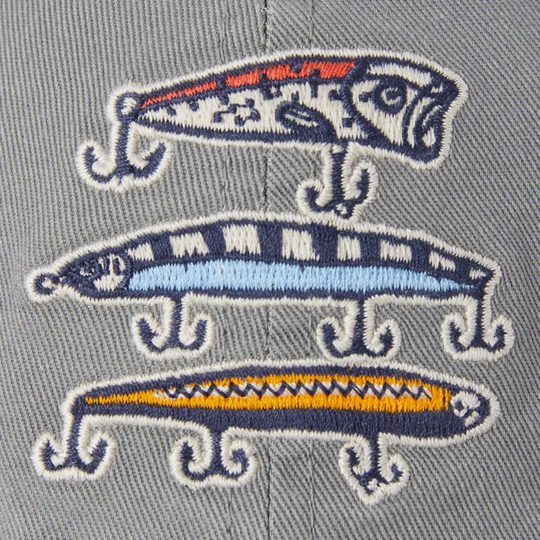 Close-up of embroidered fishing lure graphics on a gray fabric background. The embroidery features three different lures in navy blue, light blue, orange, and yellow with white outlines.