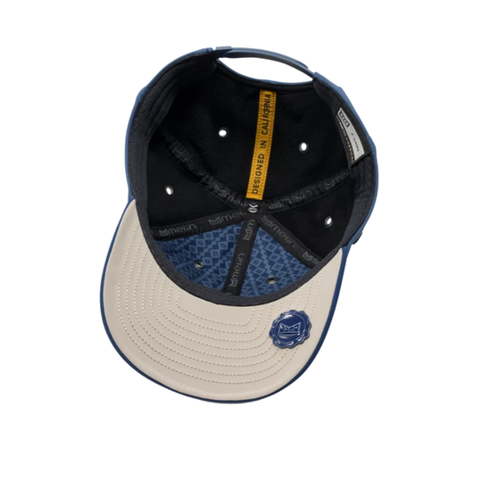 An overhead view of a baseball cap with a cream-colored curved brim, navy blue trim, a navy blue patterned underbrim, and a black interior. A small blue circular logo is visible on the front of the brim.
