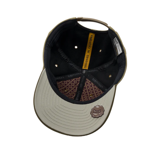 An overhead view of a baseball cap with a light beige curved brim, black interior, and brown patterned lining, featuring a small metallic rose emblem on the brim.