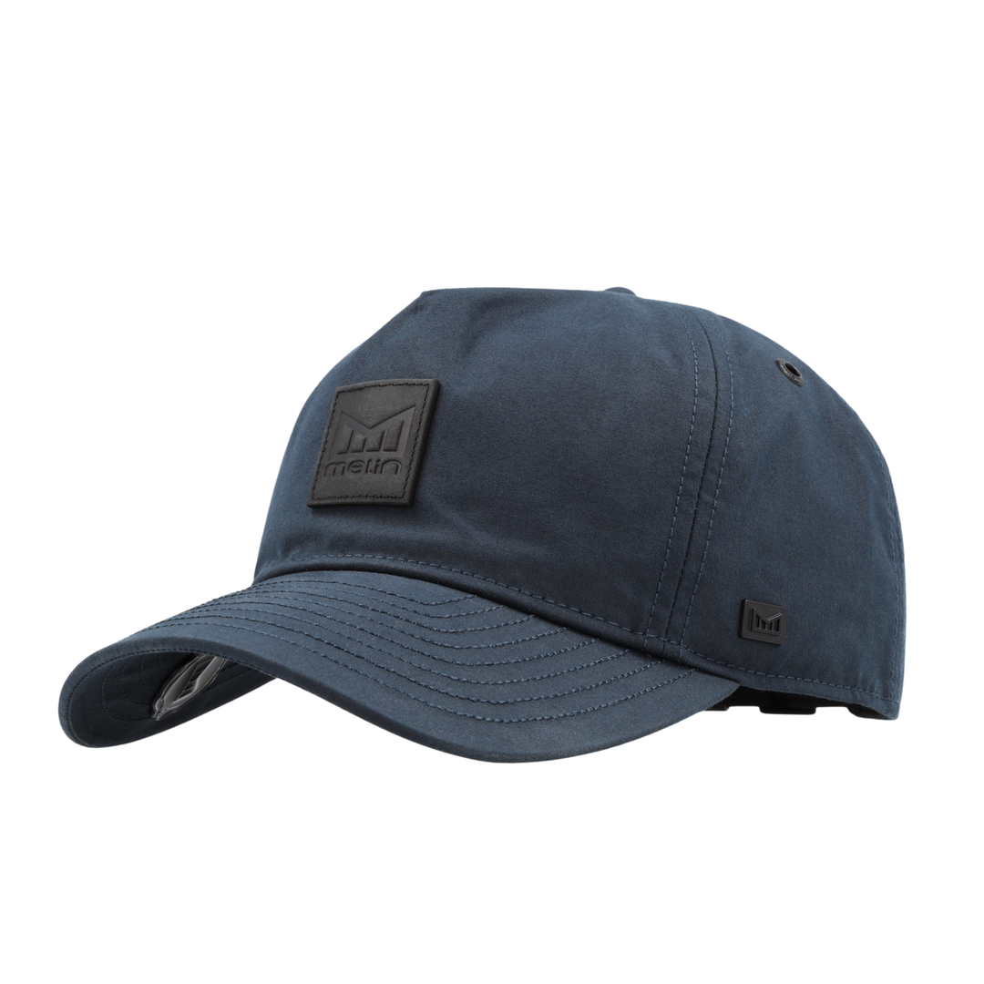 A navy blue baseball cap with a curved brim, featuring a black rectangular logo patch on the front and a small black logo on the side, set against a plain white background.