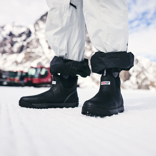 Black waterproof snow boots with rolled cuffs worn with white snow pants in a snowy mountain setting.