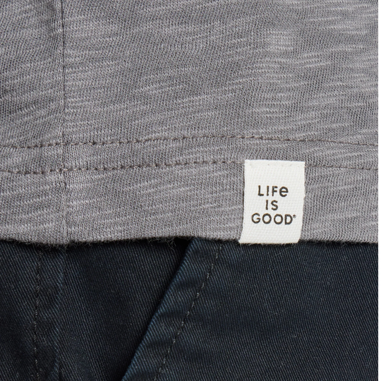 Close-up of a gray heather knit garment with a white fabric tag reading 'LIFE IS GOOD' near the hem, paired with dark navy pants.