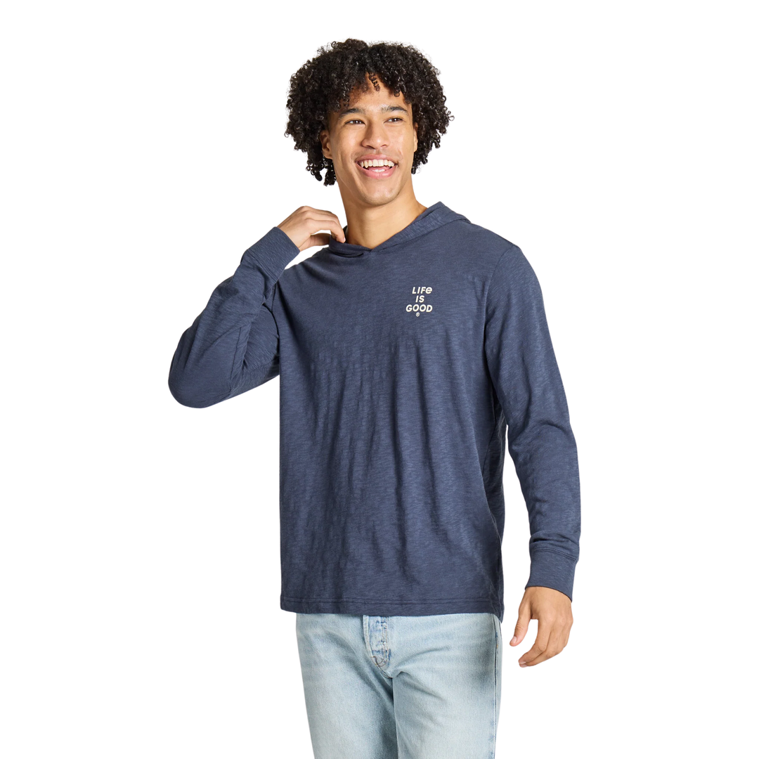 A smiling man with curly dark hair wearing a heathered navy blue long-sleeve t-shirt with a small white graphic on the left chest, paired with light blue jeans, against a solid white background.