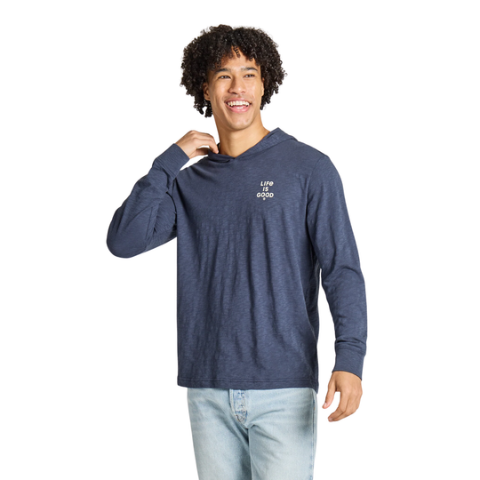 A smiling man with curly dark hair wearing a heathered navy blue long-sleeve t-shirt with a small white graphic on the left chest, paired with light blue jeans, against a solid white background.