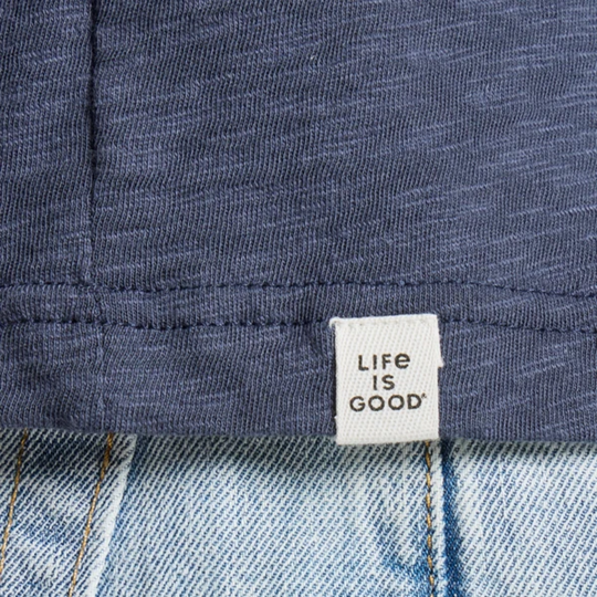 Close-up of a dark heathered blue fabric garment with a white woven label reading 'LIFE IS GOOD' placed over light blue denim.