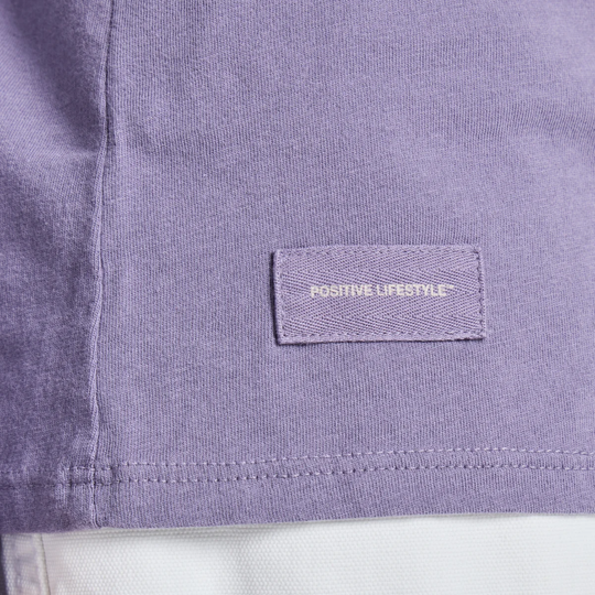 Close-up of a lavender-purple t-shirt showing a stitched rectangular brand patch that reads 'POSITIVE LIFESTYLE' in white text.