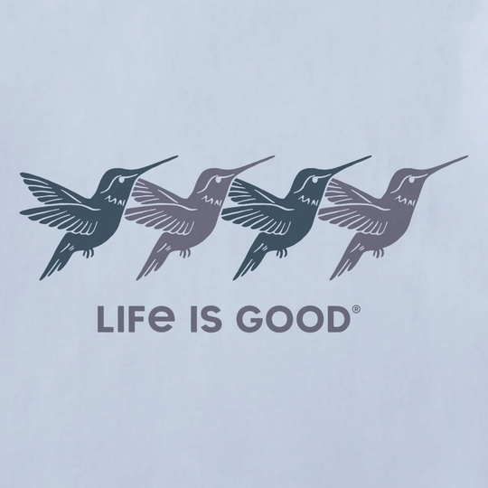 A close-up of a light blue garment featuring a printed graphic of four hummingbirds in dark gray and muted purple shades above the text 'LIFE IS GOOD' in dark gray.
