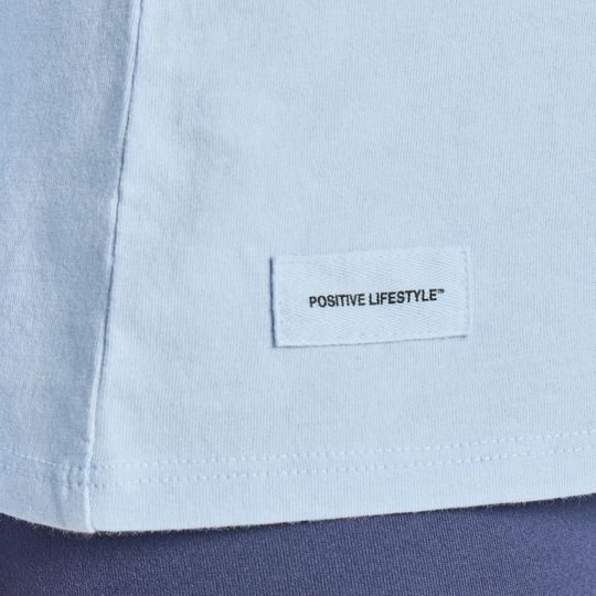 Close-up of a light blue t-shirt showing the hem with a white rectangular label that reads 'POSITIVE LIFESTYLE' in black text, layered over a dark navy garment.