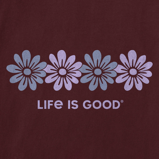 A close-up of a dark maroon or burgundy fabric with a printed graphic of four stylized flowers in alternating light blue and pale lavender, above the text 'LIFE IS GOOD' in matching light lavender.