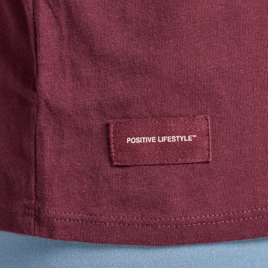 Close-up of a maroon knit garment with a rectangular sewn label reading “POSITIVE LIFESTYLE™” in white, shown against a light blue fabric background.
