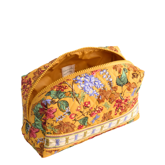 A rectangular zippered cosmetic pouch with a mustard yellow background and a detailed botanical print featuring blue flowers, green leaves, and red berries. The pouch is shown open, revealing a gold-toned interior lining and a decorative embroidered band along the bottom zipper.