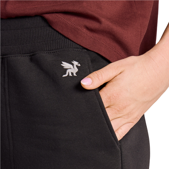 Close-up of a person wearing black sweatpants with a side pocket, showing a hand in the pocket and a small embroidered winged logo on the pocket. A burgundy top is also visible at the top of the frame against a white background.