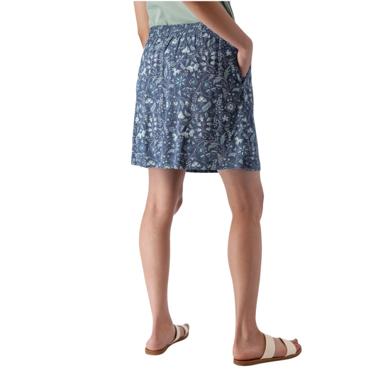 A person shown from the back wearing a blue patterned skirt with side pockets, paired with a light green top and beige slide sandals, against a plain white background.