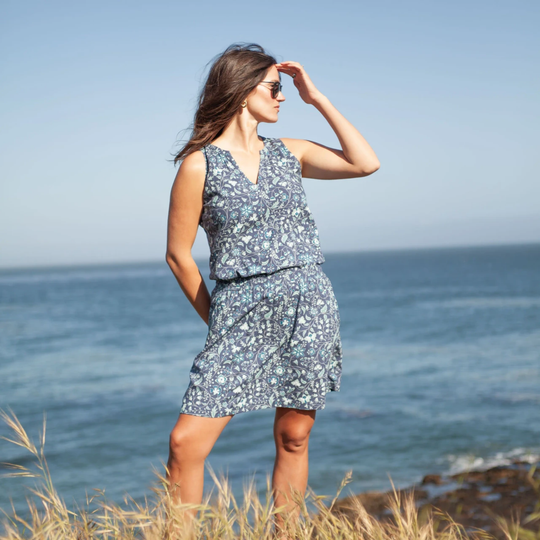 A woman wearing a sleeveless V-neck romper with a blue and white floral pattern stands on a grassy cliff overlooking the ocean.