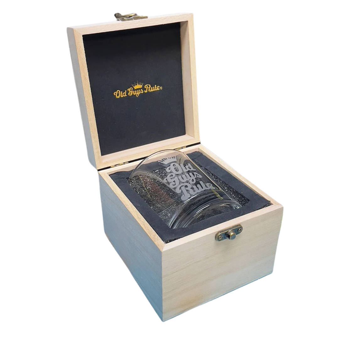 A clear glass tumbler with 'Old Guys  Rule' printed in white, presented inside an open light wood gift box with a black foam insert and gold logo on the inside lid.
