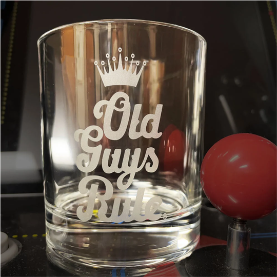 A clear glass rocks tumbler with a white frosted etched design that reads 'Old Guys Rule' under a small crown, shown on a dark surface next to a red ball control.