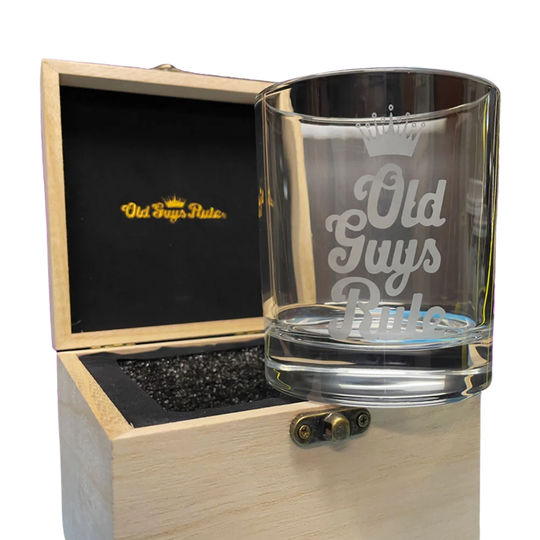 A clear engraved whiskey glass with the words 'Old Guys Rule' and a crown graphic, displayed next to an open light wood presentation box with a black interior and gold logo.