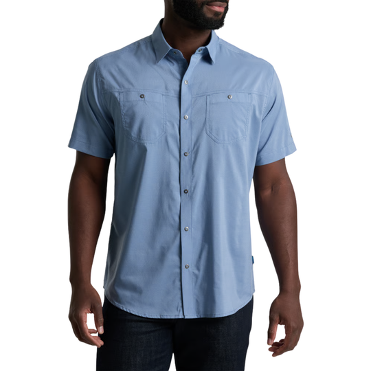 A man wearing a light blue short-sleeve button-up shirt with two chest pockets, paired with dark jeans, shown against a plain white background.