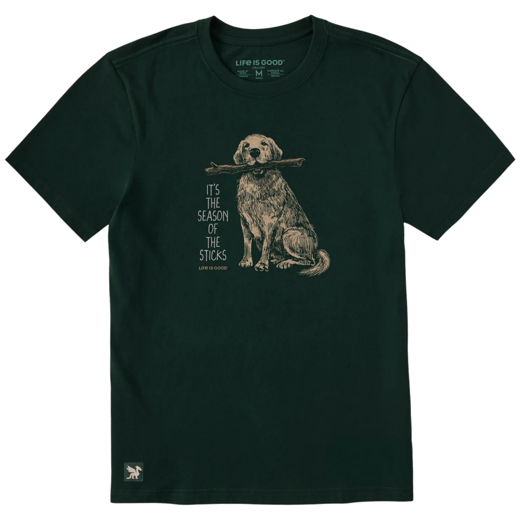 A dark forest green short-sleeve t-shirt with a graphic of a golden retriever holding a stick, next to the text 'IT'S THE SEASON OF THE STICKS'. The brand 'LIFE IS GOOD' is visible on the inner neck label and as a small logo on the bottom left hem.