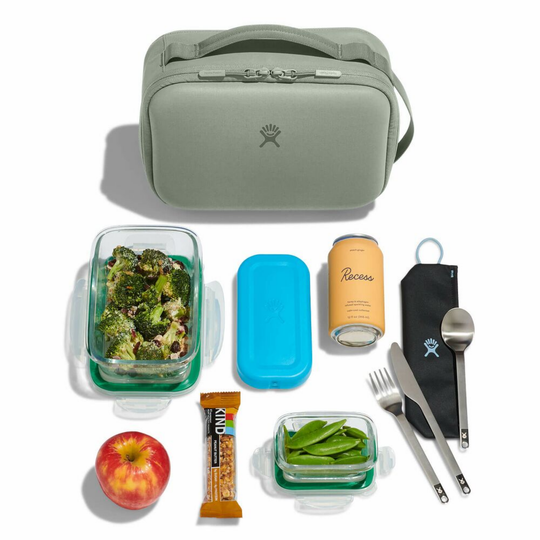 Green lunch bag with food containers, apple, snack bar, and utensils on a white background