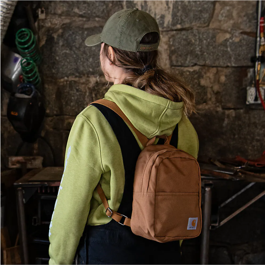 A person wearing a light green hoodie, olive green cap, and black top carries a tan Carhartt canvas backpack with brown straps in a rustic workshop setting.