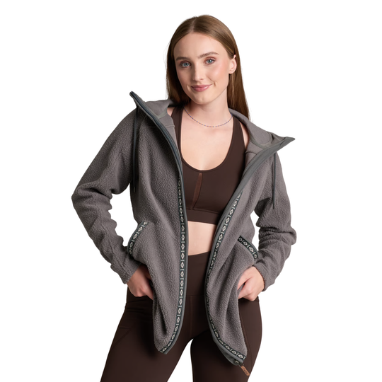 A woman modeling a gray fleece zip-up hoodie with dark green patterned zipper trim, worn open over a brown sports bra and matching brown leggings, against a plain white background.