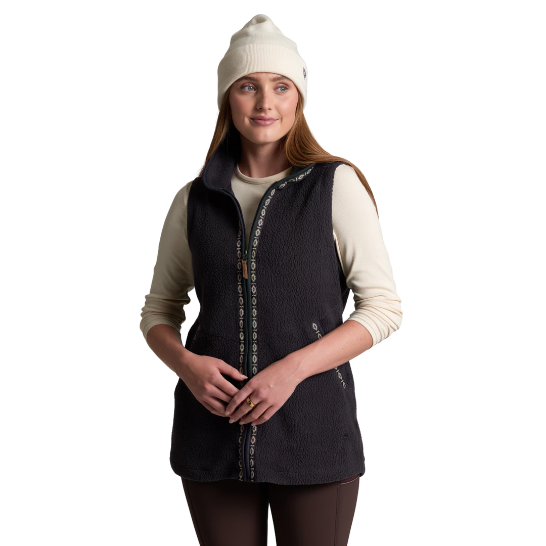A woman wearing a dark charcoal fleece vest over a cream long-sleeve shirt, paired with a cream beanie and dark leggings, standing against a solid white background.