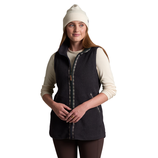 A woman wearing a dark charcoal fleece vest over a cream long-sleeve shirt, paired with a cream beanie and dark leggings, standing against a solid white background.