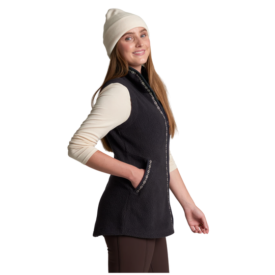 A woman wearing a sleeveless black fleece vest over a cream long-sleeve shirt, paired with a cream beanie and brown leggings, shown against a plain white background.