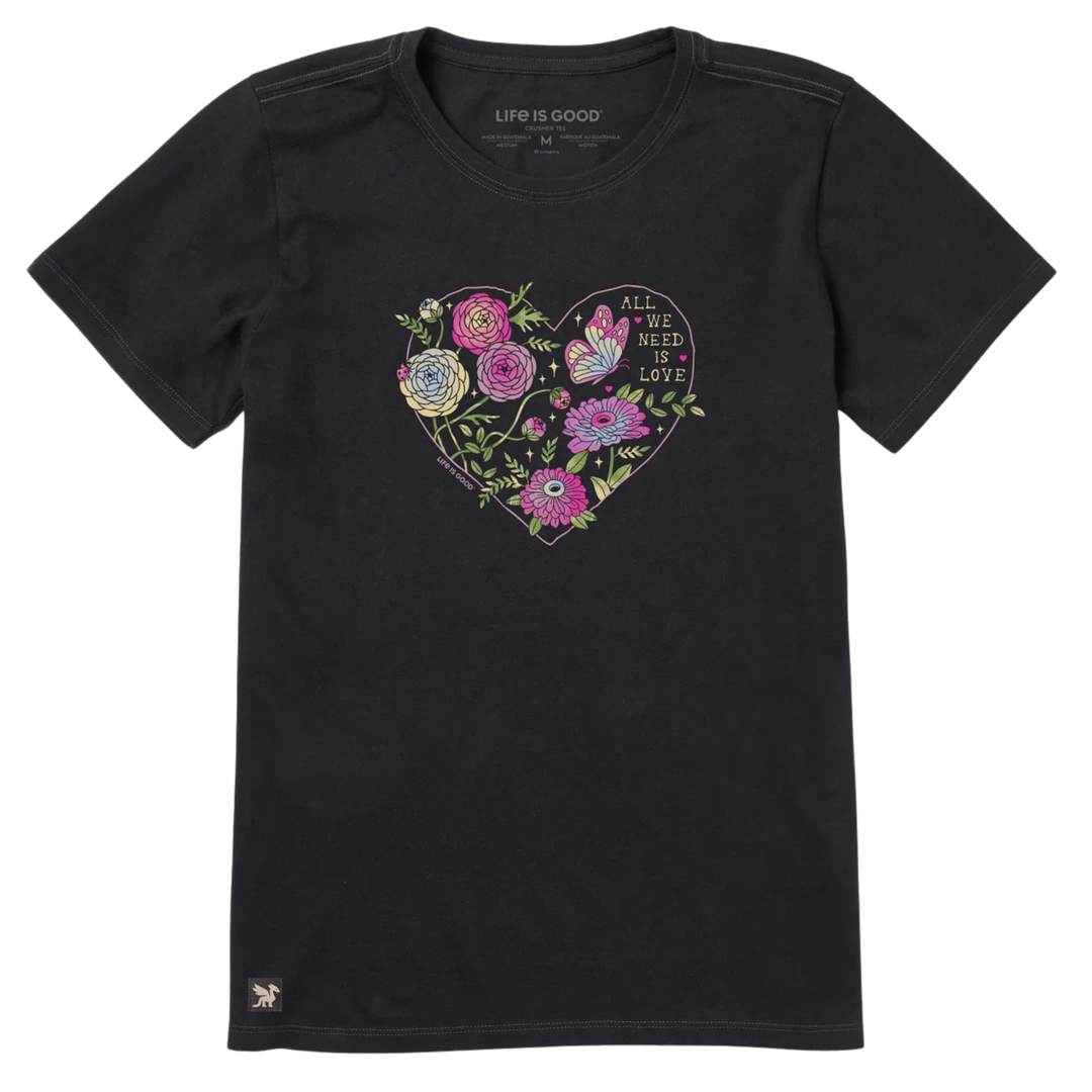 A black short-sleeve crewneck t-shirt featuring a colorful heart-shaped graphic on the chest with flowers, a butterfly, and the text 'ALL WE NEED IS LOVE'. The brand name 'LIFE IS GOOD' is printed inside the collar, and a small logo is visible near the bottom left hem.