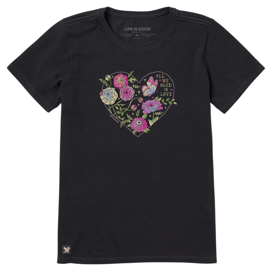 A black short-sleeve crewneck t-shirt featuring a colorful heart-shaped graphic on the chest with flowers, a butterfly, and the text 'ALL WE NEED IS LOVE'. The brand name 'LIFE IS GOOD' is printed inside the collar, and a small logo is visible near the bottom left hem.