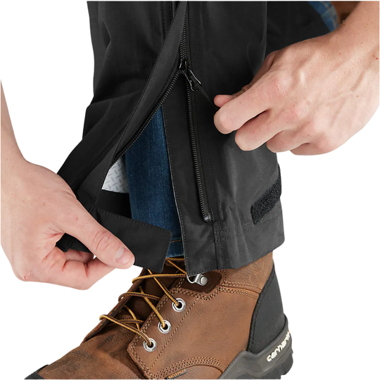 Close-up of a person's hands unzipping and pulling down the cuff of black waterproof pants, showing a brown Carhartt work boot and blue jeans inside. The Carhartt brand is visible on the heel of the boot.