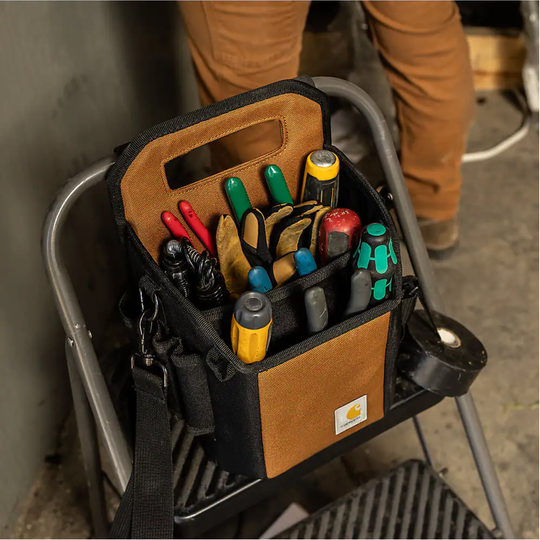 A brown and black Carhartt tool bag filled with hand tools, resting on a metal step ladder in a workshop setting.
