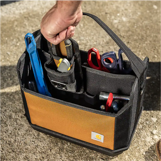 A black and tan Carhartt tool caddy bag filled with hand tools, held by a person's hand outdoors on a concrete surface.