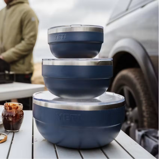 Three nested YETI food storage bowls in a dark slate blue color with stainless steel rims, stacked on a white slatted table outdoors with a car and a person blurred in the background.