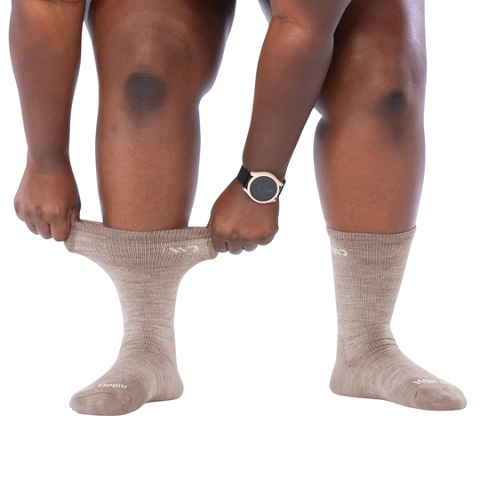 A person wearing ribbed ankle socks in a light taupe-beige color, pulling one sock up with both hands to demonstrate elasticity. The person is also wearing a black and silver smartwatch on their wrist, set against a solid white background.