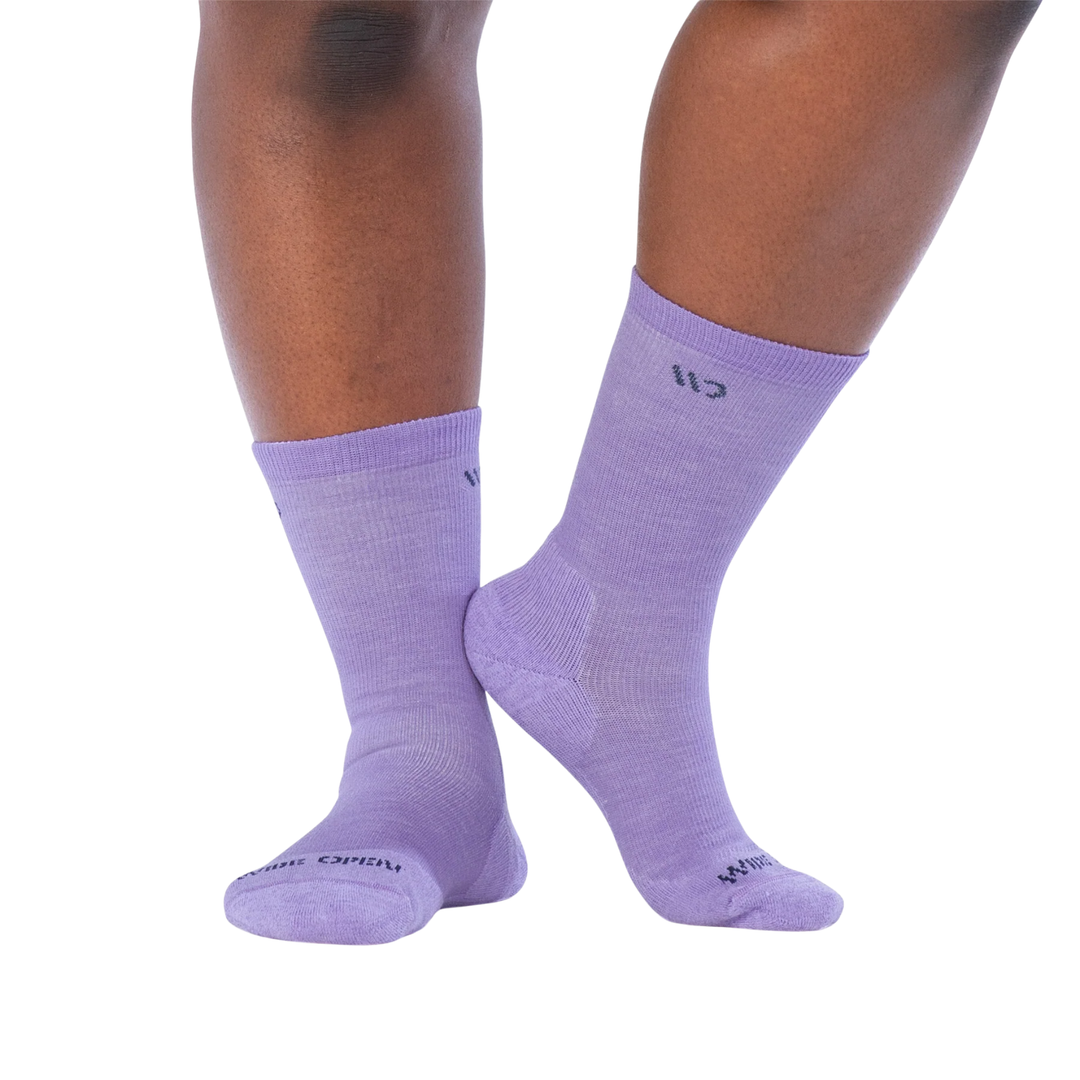 A pair of light purple crew socks worn on a person's feet, shown against a plain white background. The socks feature a small dark logo on the upper calf and text near the toe.