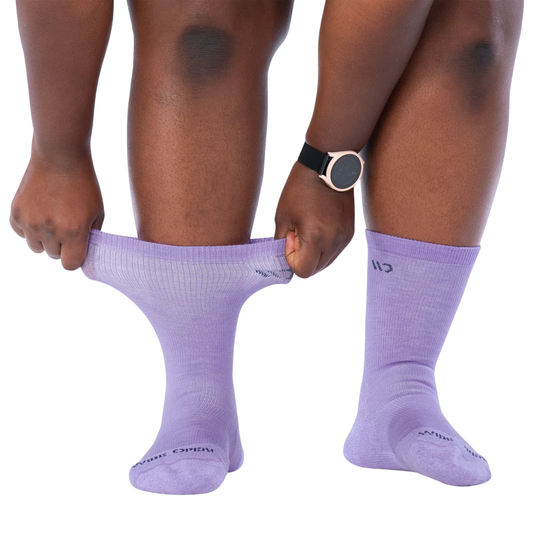 A person wearing light purple crew socks, pulling one sock up with both hands to show flexibility and fit. The person has a black and rose gold smartwatch on their wrist.