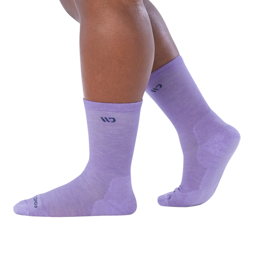 A pair of light purple crew socks worn on a person's feet, shown against a plain white background. The socks feature a small dark logo on the upper calf and text near the toe.