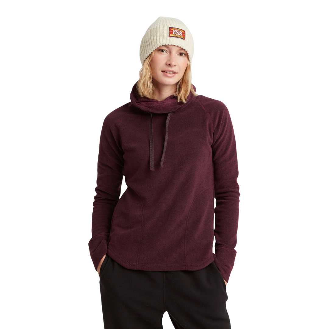 A woman wearing a dark burgundy cowl-neck long-sleeve fleece pullover, black sweatpants, and a cream-colored knit beanie with a small orange logo patch. She is standing against a solid white background.