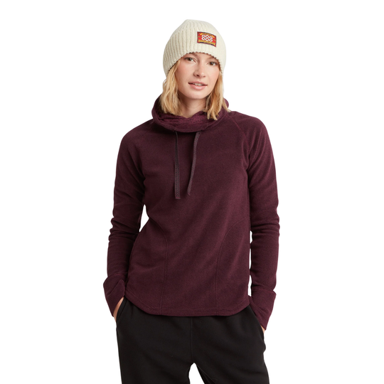 A woman wearing a dark burgundy cowl-neck long-sleeve fleece pullover, black sweatpants, and a cream-colored knit beanie with a small orange logo patch. She is standing against a solid white background.
