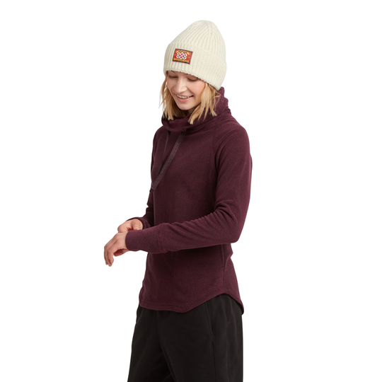 A woman wearing a burgundy long-sleeve mock neck fleece, a cream knit beanie with a small orange and white logo patch, and black pants, photographed against a solid white background.
