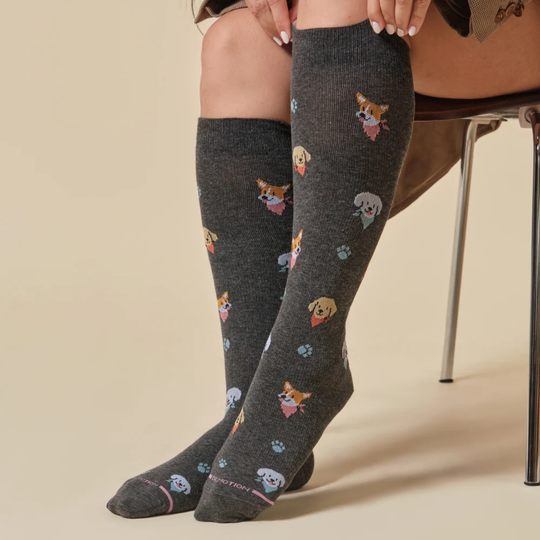 A person seated on a chair wearing dark charcoal knee-high socks with a playful pattern of small dog faces, paw prints, and tiny colorful accents. The photo is cropped from the waist down against a warm beige studio background.