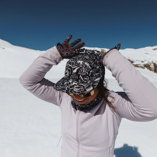 A woman in a snowy mountain setting wearing a black and white floral patterned winter hat with a matching neck gaiter, black gloves with red accents, and a light pink long-sleeve jacket.